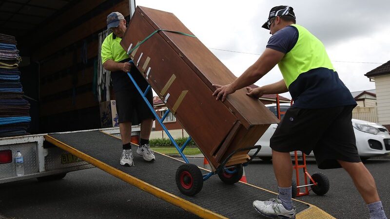 Two movers wheeling a large strapped cabinet on a hand truck up a ramp into a removal truck