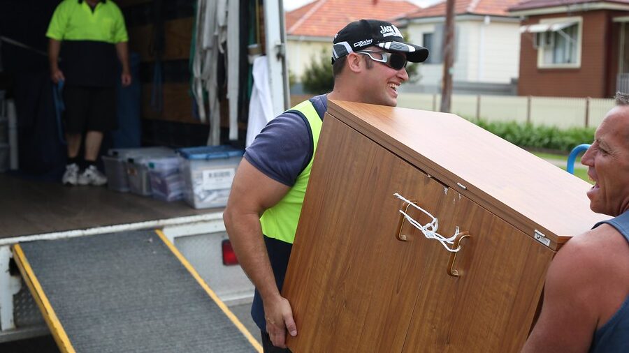 Removalists carrying a wooden cabinet toward an open removal truck with a loading ramp on a suburban street