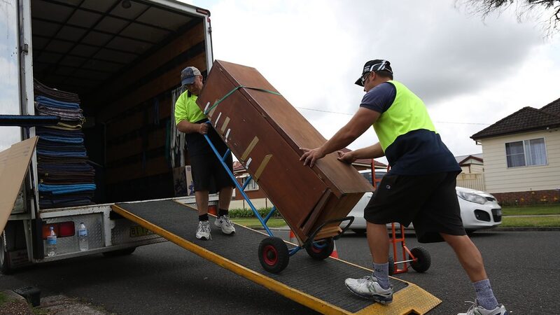 Two removalists in hi-vis vests wheeling a large wooden cabinet up a ramp into a removal truck with a hand truck