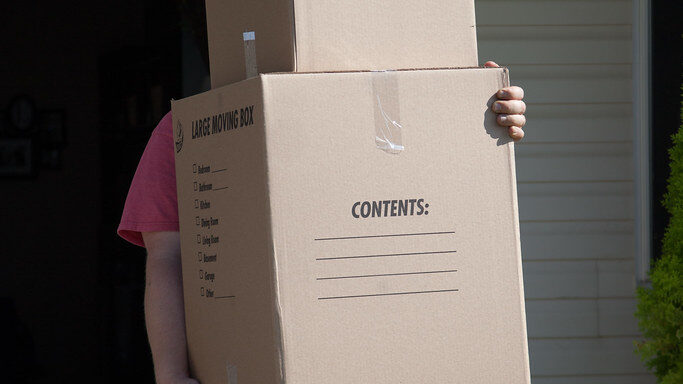 Person in jeans carrying a large labelled cardboard moving box outside a house on moving day in Christchurch