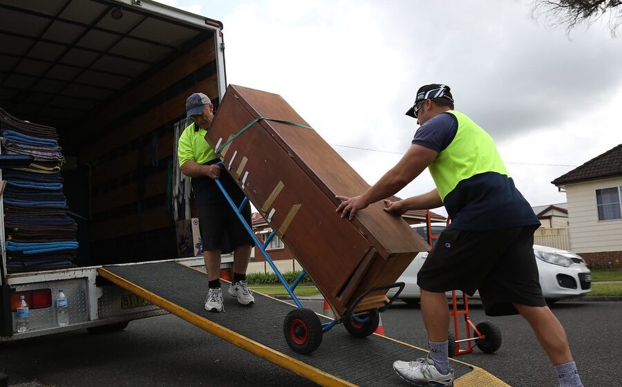 Movers guiding a strapped wooden cabinet on a hand truck up the loading ramp of a removal truck