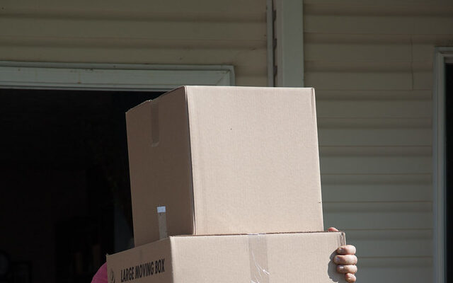 Removalist carrying stacked moving boxes labelled with contents outside a suburban home