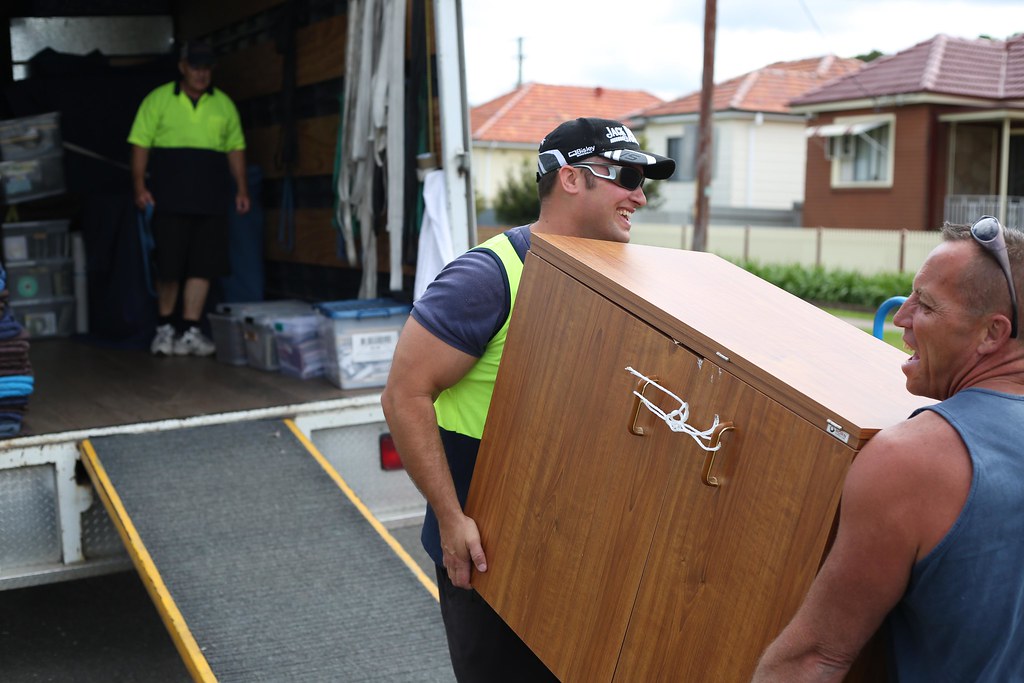 Two removalists in hi-vis vests carrying a wooden cabinet up a ramp into a removal truck on a suburban street