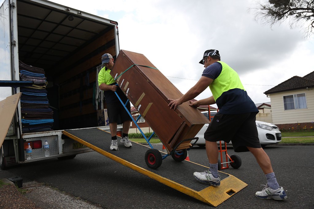 Two removalists in hi-vis vests wheeling a wooden cabinet up a ramp into a removal truck on a suburban street