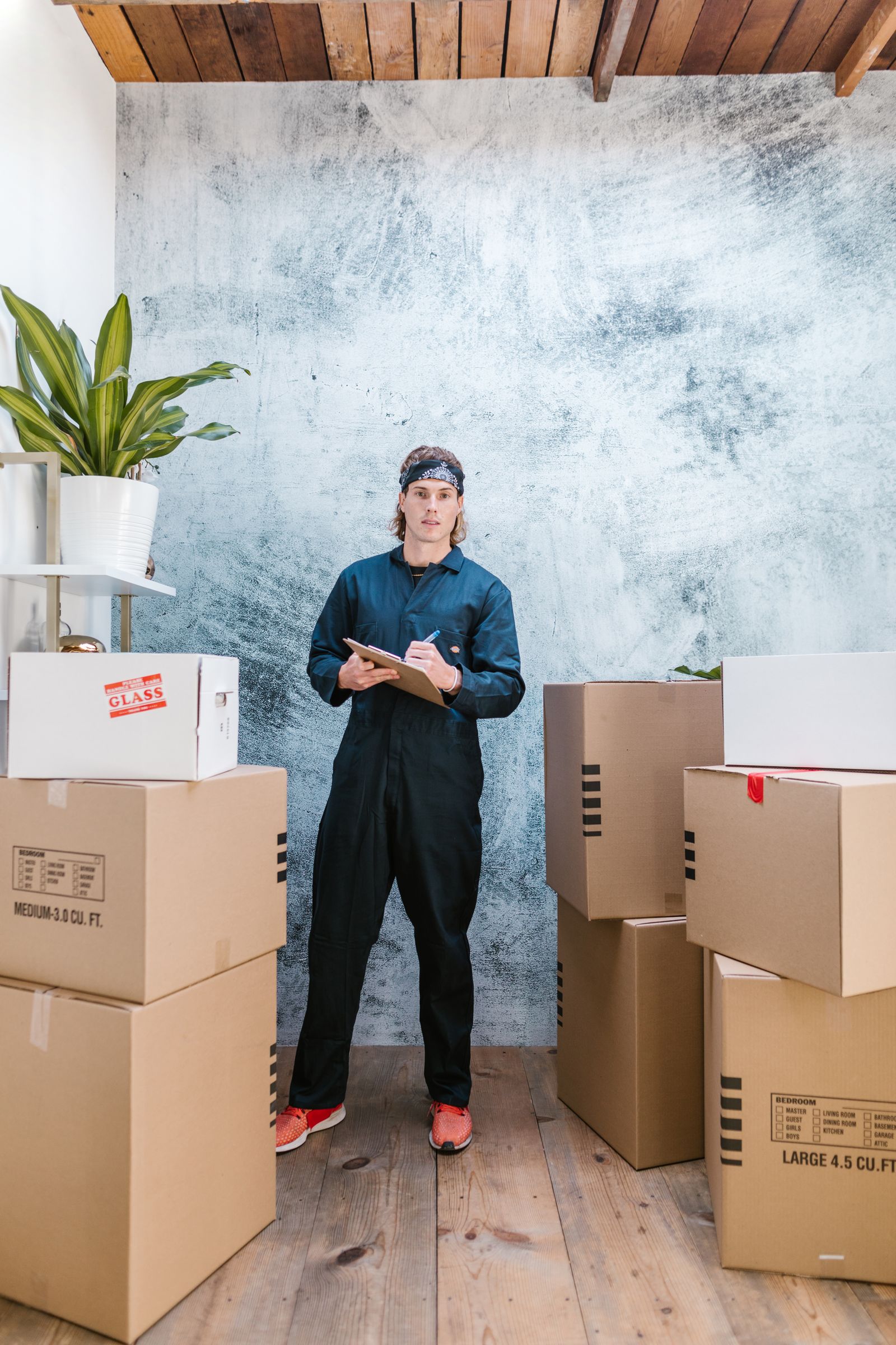 Mover in coveralls writing on a clipboard surrounded by packed moving boxes